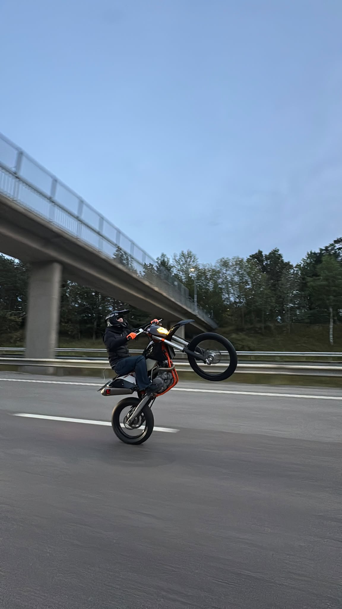 Rider wheelieing an orange-framed supermoto beneath a highway overpass