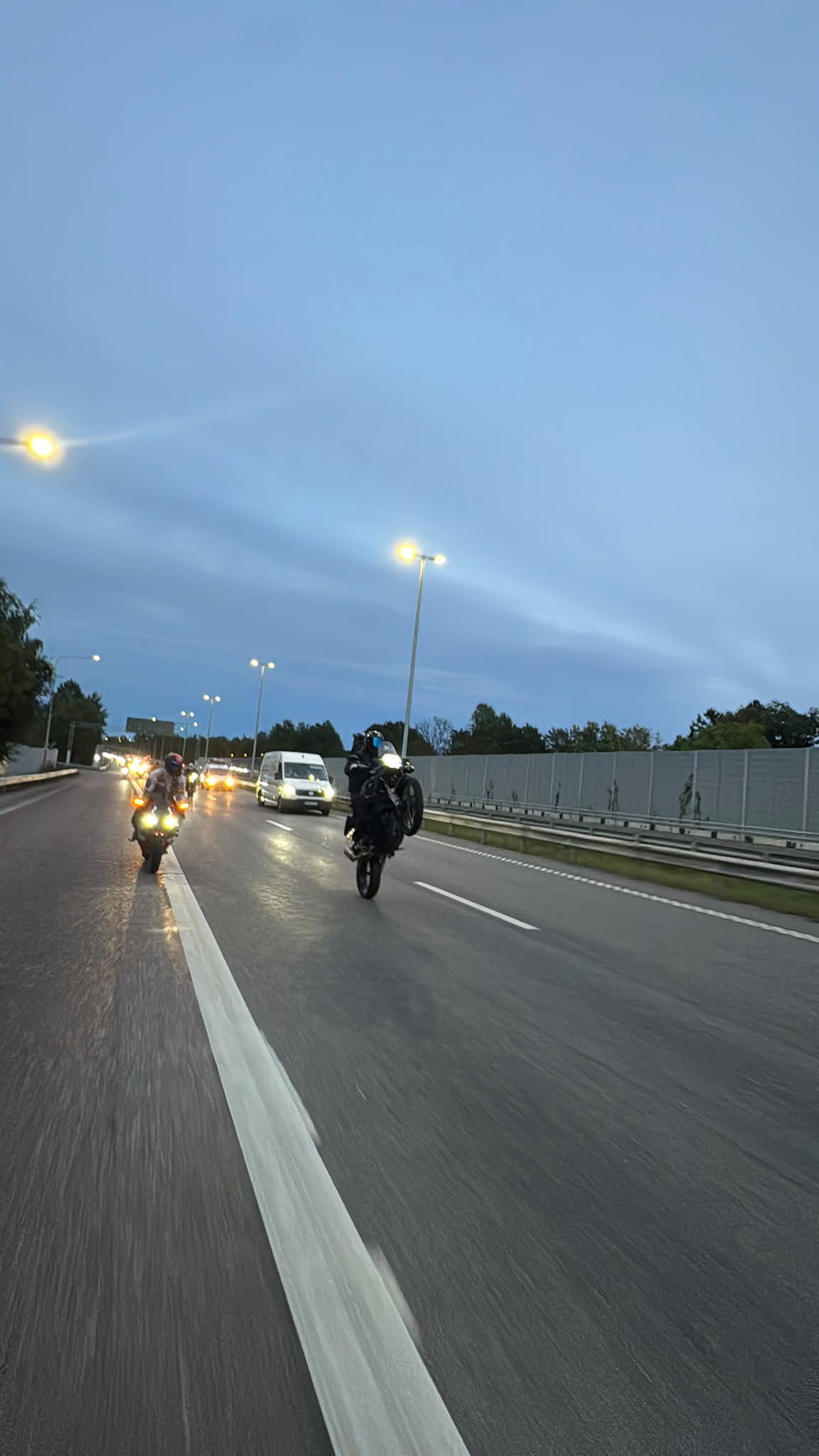 Riders with headlights on at dusk, one lifting the front wheel under street lights